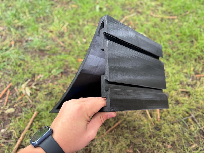May include: A black, angular object with a ridged design, held in a hand. The object has a triangular shape with horizontal grooves. The background is blurred green grass and foliage.
