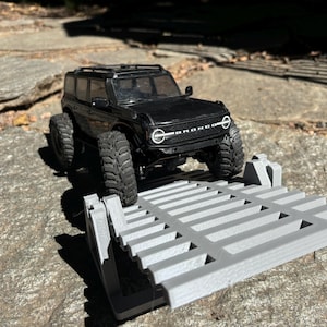 K&ouml;nnte beinhalten: Ein schwarzes Spielzeug-Bronco-SUV mit gro&szlig;en Reifen steht auf einer grauen, 3D-gedruckten Rampe. Das Fahrzeug hat einen Dachgep&auml;cktr&auml;ger und wei&szlig;e Scheinwerfer. Die Szene spielt sich im Freien auf einer Steinoberfl&auml;che ab.
