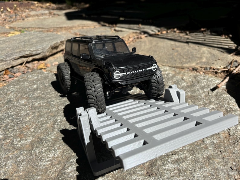 K&ouml;nnte beinhalten: Ein schwarzes Spielzeug-Bronco-SUV mit gro&szlig;en Reifen steht auf einer grauen, 3D-gedruckten Rampe. Das Fahrzeug hat einen Dachgep&auml;cktr&auml;ger und wei&szlig;e Scheinwerfer. Die Szene spielt sich im Freien auf einer Steinoberfl&auml;che ab.