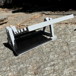 K&ouml;nnte beinhalten: Ein hellgraues 3D-gedrucktes Objekt, m&ouml;glicherweise ein Werkzeug oder eine Halterung, mit rechteckiger Basis und abgewinkeltem Arm. Das Objekt liegt auf einer strukturierten, grauen Steinoberfl&auml;che, mit Schatten.