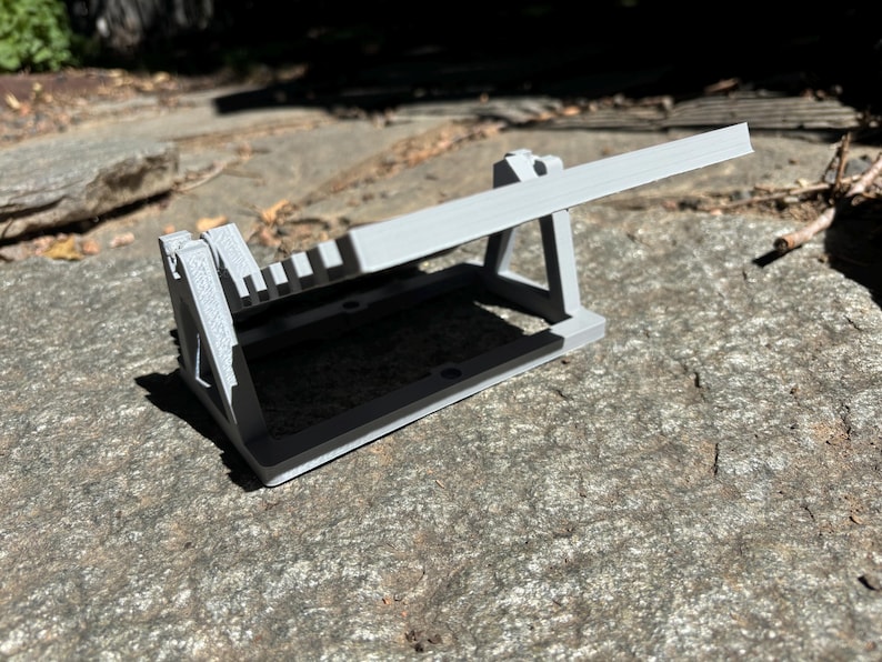 K&ouml;nnte beinhalten: Ein hellgraues 3D-gedrucktes Objekt, m&ouml;glicherweise ein Werkzeug oder eine Halterung, mit rechteckiger Basis und abgewinkeltem Arm. Das Objekt liegt auf einer strukturierten, grauen Steinoberfl&auml;che, mit Schatten.