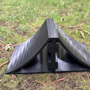May include: A black, angular object with a ridged surface, resembling a tent or shelter, is positioned on a grassy area. The object appears to be made of a solid material and has a curved base. The design suggests a potential for outdoor use.