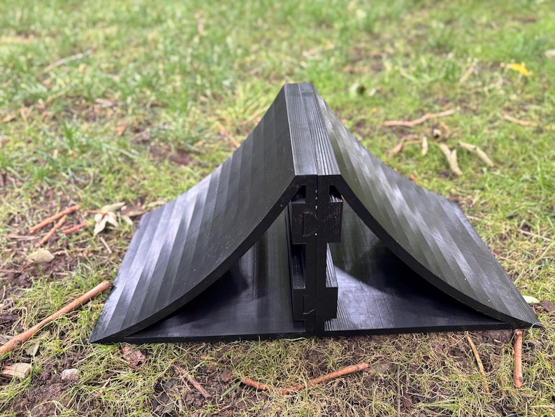 May include: A black, angular object with a ridged surface, resembling a tent or shelter, is positioned on a grassy area. The object appears to be made of a solid material and has a curved base. The design suggests a potential for outdoor use.