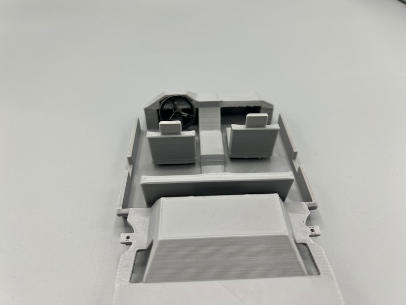 May include: A light gray 3D-printed model of a vehicle interior. The model includes a steering wheel, two seats, and a dashboard. The interior is a simplified representation of a car's cockpit.