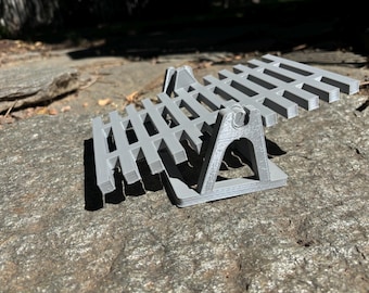 Könnte beinhalten: Ein grauer, 3D-gedruckter, zweistöckiger Ständer mit Lamellendesign. Der Ständer wird von abgewinkelten Beinen getragen und ist zur Aufnahme eines Gegenstandes bestimmt. Das Objekt liegt auf einem Felsen.