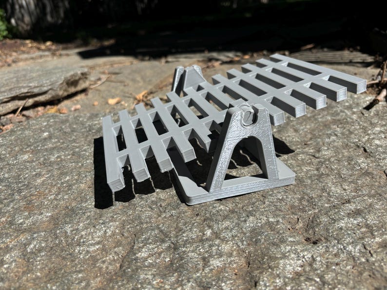 K&ouml;nnte beinhalten: Ein grauer, 3D-gedruckter, zweist&ouml;ckiger St&auml;nder mit Lamellendesign. Der St&auml;nder wird von abgewinkelten Beinen getragen und ist zur Aufnahme eines Gegenstandes bestimmt. Das Objekt liegt auf einem Felsen.