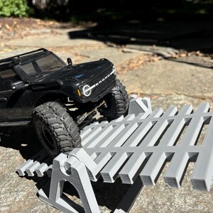 K&ouml;nnte beinhalten: Ein schwarzes Spielzeug-SUV mit gro&szlig;en Reifen steht auf einer grauen, 3D-gedruckten Rampenstruktur. Das Fahrzeug ist abgewinkelt, was darauf hindeutet, dass es die Rampe bef&auml;hrt. Die Szene spielt im Freien an einem sonnigen Tag.