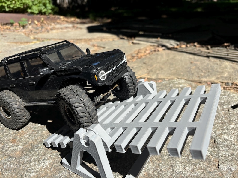 K&ouml;nnte beinhalten: Ein schwarzes Spielzeug-SUV mit gro&szlig;en Reifen steht auf einer grauen, 3D-gedruckten Rampenstruktur. Das Fahrzeug ist abgewinkelt, was darauf hindeutet, dass es die Rampe bef&auml;hrt. Die Szene spielt im Freien an einem sonnigen Tag.