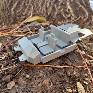 May include: A light gray 3D-printed model of a vehicle interior, featuring seats and a dashboard. The model is resting on a bed of soil and leaves, with a tree trunk in the background. The model appears to be a miniature representation of a car's interior.