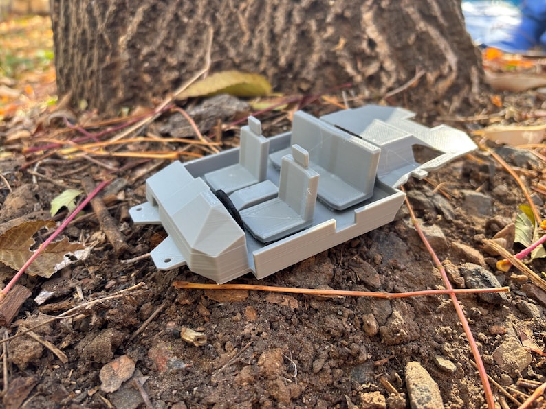 May include: A light gray 3D-printed model of a vehicle interior, featuring seats and a dashboard. The model is resting on a bed of soil and leaves, with a tree trunk in the background. The model appears to be a miniature representation of a car's interior.