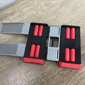 May include: A black and gray 3D-printed device with red cylindrical objects. The device has two gray, perforated panels extending from the sides. The device is designed to hold multiple red cylindrical objects.