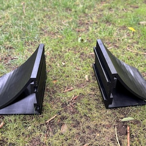 May include: Two black, wedge-shaped objects with a curved design and ribbed detailing. The objects are made of a matte material and are positioned on a bed of green grass. The objects appear to be a pair.