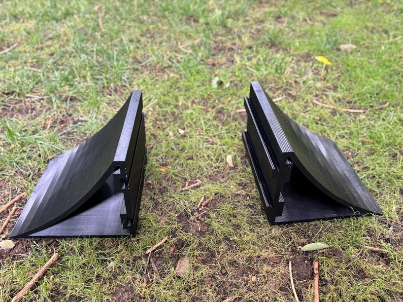May include: Two black, wedge-shaped objects with a curved design and ribbed detailing. The objects are made of a matte material and are positioned on a bed of green grass. The objects appear to be a pair.