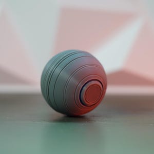 K&ouml;nnte beinhalten: Eine grau-rote Kugel mit konzentrischen Rillen und einem zentralen kreisf&ouml;rmigen Design. Das Objekt scheint ein Stressball oder ein dekorativer Gegenstand zu sein. Die Kugel liegt auf einer dunklen Oberfl&auml;che.
