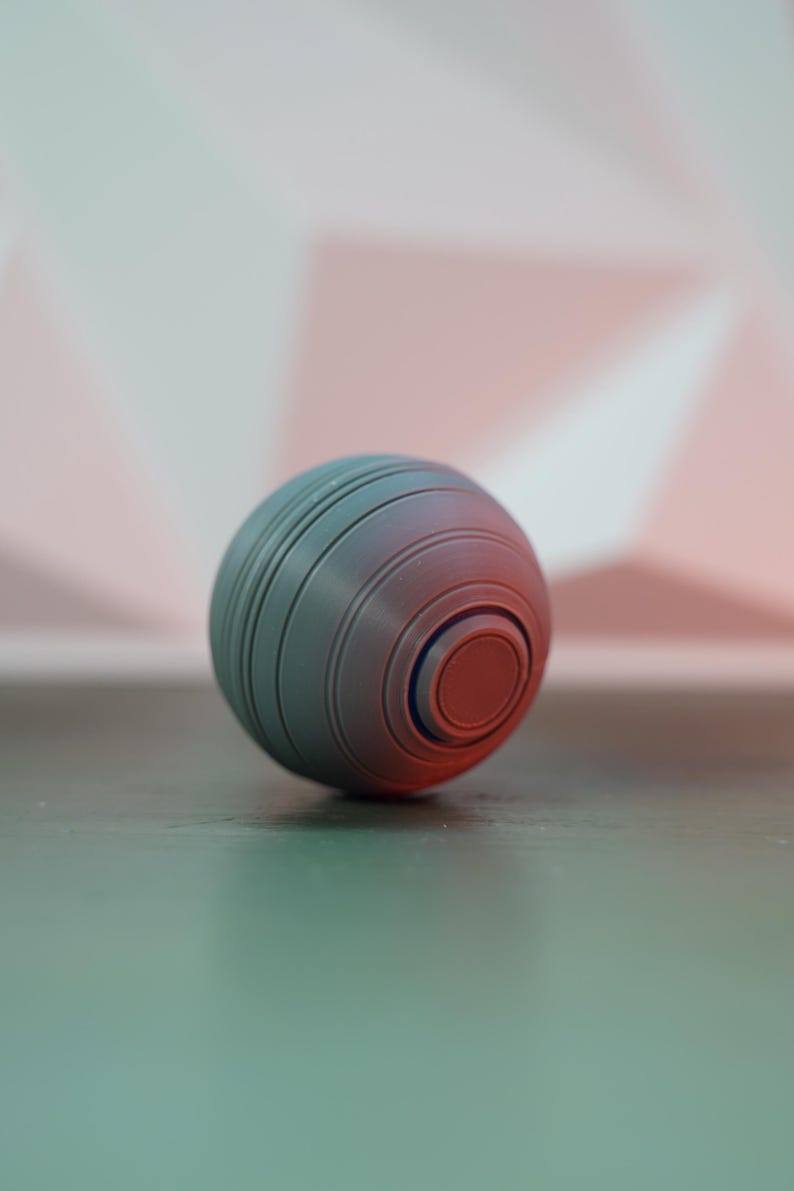 K&ouml;nnte beinhalten: Eine grau-rote Kugel mit konzentrischen Rillen und einem zentralen kreisf&ouml;rmigen Design. Das Objekt scheint ein Stressball oder ein dekorativer Gegenstand zu sein. Die Kugel liegt auf einer dunklen Oberfl&auml;che.