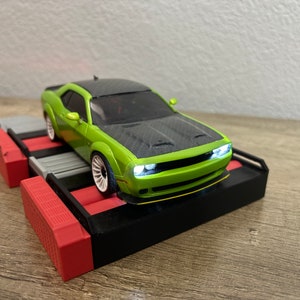 May include: A green and black toy car with white wheels and working headlights sits on a black, red, and gray platform. The car has a carbon fiber hood and a yellow accent on the front bumper. The platform has a red and gray design.
