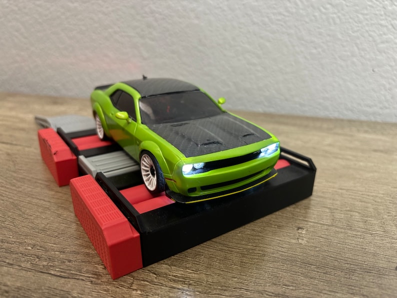 May include: A green and black toy car with white wheels and working headlights sits on a black, red, and gray platform. The car has a carbon fiber hood and a yellow accent on the front bumper. The platform has a red and gray design.