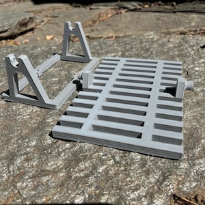 K&ouml;nnte beinhalten: Ein hellgraues 3D-gedrucktes Set von Objekten, darunter ein rechteckiger Rost mit horizontalen Schlitzen und zwei abgewinkelten St&uuml;tzstrukturen. Die Objekte bestehen aus Kunststoff und sind so konzipiert, dass sie zusammenpassen. Das Set wird auf einer strukturierten Steinoberfl&auml;che pr&auml;sentiert.