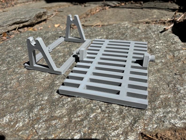 K&ouml;nnte beinhalten: Ein hellgraues 3D-gedrucktes Set von Objekten, darunter ein rechteckiger Rost mit horizontalen Schlitzen und zwei abgewinkelten St&uuml;tzstrukturen. Die Objekte bestehen aus Kunststoff und sind so konzipiert, dass sie zusammenpassen. Das Set wird auf einer strukturierten Steinoberfl&auml;che pr&auml;sentiert.