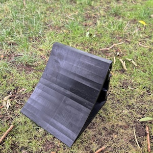 May include: A black wheel chock, designed to prevent vehicle movement, rests on a bed of grass. The chock has a ridged surface and a wedge shape, ideal for securing tires on inclines or during maintenance.