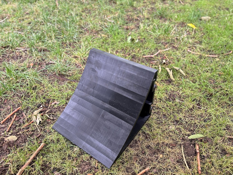 May include: A black wheel chock, designed to prevent vehicle movement, rests on a bed of grass. The chock has a ridged surface and a wedge shape, ideal for securing tires on inclines or during maintenance.