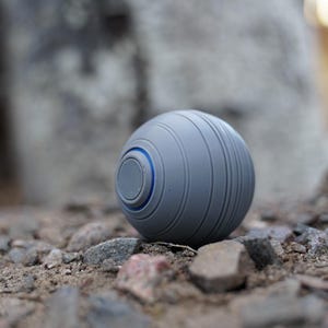 K&ouml;nnte beinhalten: Ein grauer Ball mit einem blauen Akzentring, der auf einem Bett aus kleinen Steinen ruht. Der Ball hat eine Reihe paralleler Rillen um seinen Umfang. Das Design ist modern und minimalistisch.