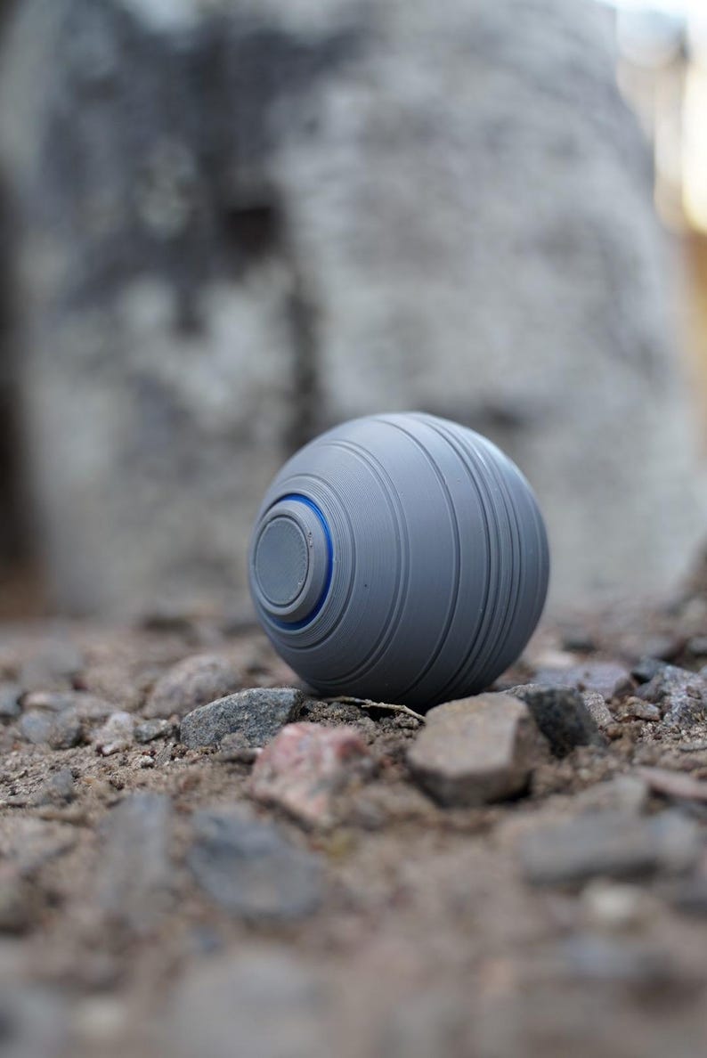 K&ouml;nnte beinhalten: Ein grauer Ball mit einem blauen Akzentring, der auf einem Bett aus kleinen Steinen ruht. Der Ball hat eine Reihe paralleler Rillen um seinen Umfang. Das Design ist modern und minimalistisch.