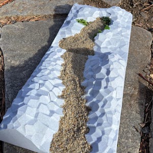 Puede incluir: Una superficie blanca texturizada con un camino elevado de arena y dos pequeñas plantas verdes. La superficie está diseñada para parecer una cordillera.