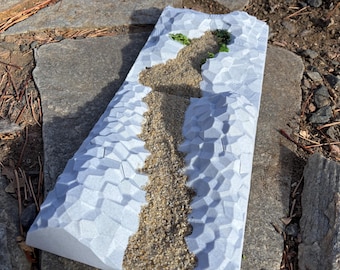 May include: A white textured surface with a raised path of sand and two small green plants. The surface is designed to look like a mountain range.