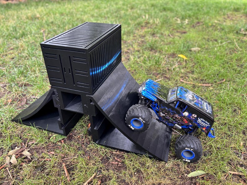 May include: A black ramp and container set for a toy monster truck. The truck is blue and black with large tires and the words "Grave Digger" on the side. The set is on a grassy surface.