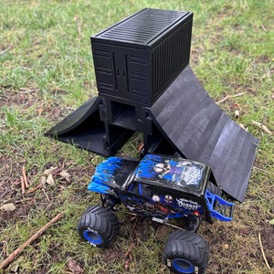 May include: A black toy cargo container and ramp set with a blue and black monster truck. The monster truck has large tires and a skull graphic. The set is on a grassy surface.