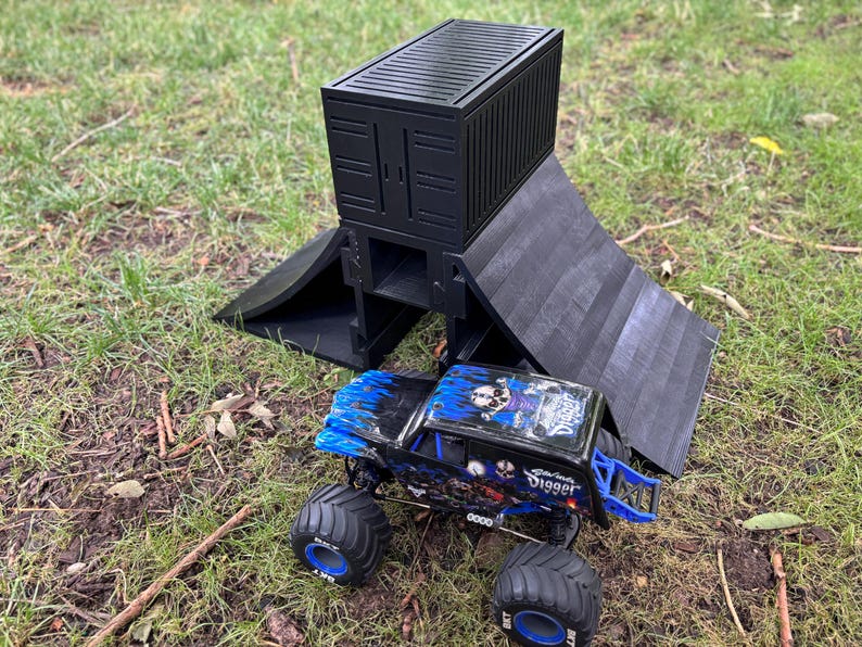 May include: A black toy cargo container and ramp set with a blue and black monster truck. The monster truck has large tires and a skull graphic. The set is on a grassy surface.