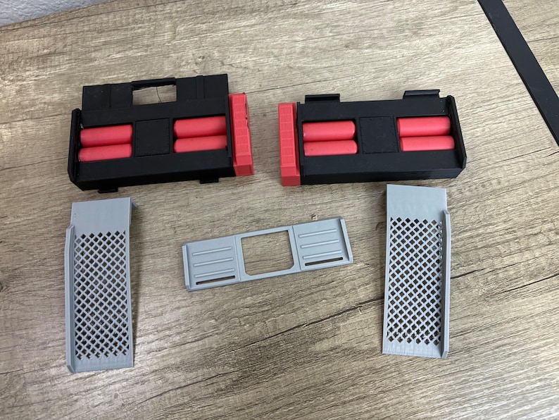 May include: An assortment of 3D-printed components in black, red, and gray. The set includes two black rectangular housings with red cylindrical inserts, two gray grilles with diamond patterns, and a gray rectangular frame.