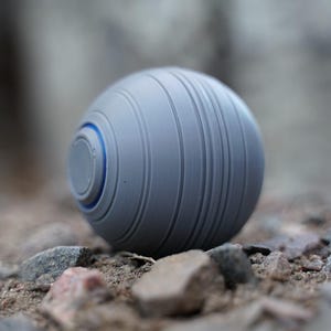 K&ouml;nnte beinhalten: Ein graues, kugelf&ouml;rmiges Objekt mit vertikalen Rillen und einem blauen Akzentring. Das Objekt liegt auf einem Bett aus kleinen Steinen und Kieselsteinen und erzeugt eine strukturierte Au&szlig;enszene.