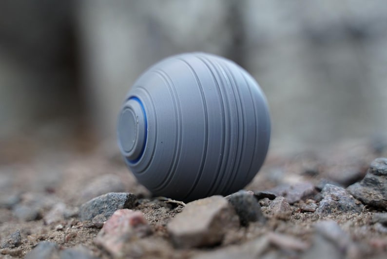 K&ouml;nnte beinhalten: Ein graues, kugelf&ouml;rmiges Objekt mit vertikalen Rillen und einem blauen Akzentring. Das Objekt liegt auf einem Bett aus kleinen Steinen und Kieselsteinen und erzeugt eine strukturierte Au&szlig;enszene.