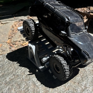 K&ouml;nnte beinhalten: Ein schwarzes Spielzeug-SUV mit gro&szlig;en, strukturierten Reifen steht auf einer grauen, abgestuften Plattform. Die gl&auml;nzende Oberfl&auml;che des Fahrzeugs reflektiert das Sonnenlicht. Die Reifen sind f&uuml;r den Gel&auml;ndeeinsatz konzipiert.