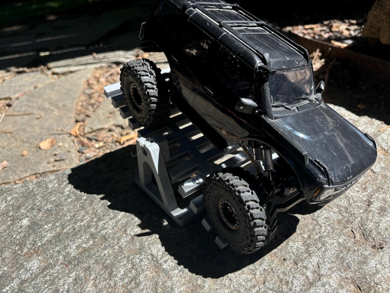 K&ouml;nnte beinhalten: Ein schwarzes Spielzeug-SUV mit gro&szlig;en, strukturierten Reifen steht auf einer grauen, abgestuften Plattform. Die gl&auml;nzende Oberfl&auml;che des Fahrzeugs reflektiert das Sonnenlicht. Die Reifen sind f&uuml;r den Gel&auml;ndeeinsatz konzipiert.