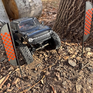 May include: A black toy truck with a red and black checkered flag on each side. The truck is on a dirt surface with rocks and sticks.