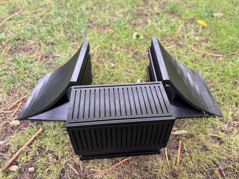 May include: Black, 3D-printed planter box with a slatted design, flanked by two matching curved supports. The planter box has a rectangular shape and is sitting on grass.