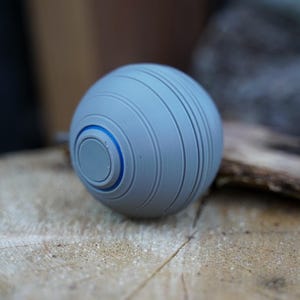 K&ouml;nnte beinhalten: Eine graue Kugel mit horizontalen Rillen und einem blauen Akzentring. Das kugelf&ouml;rmige Objekt liegt auf einer hellbraunen, strukturierten Oberfl&auml;che. Das Design ist modern und minimalistisch.