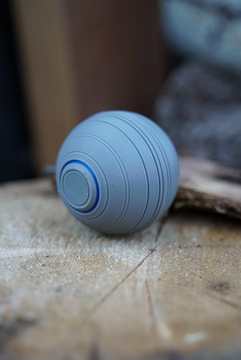 K&ouml;nnte beinhalten: Eine graue Kugel mit horizontalen Rillen und einem blauen Akzentring. Das kugelf&ouml;rmige Objekt liegt auf einer hellbraunen, strukturierten Oberfl&auml;che. Das Design ist modern und minimalistisch.