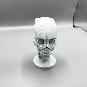 May include: A white 3D printed bust of a robot head with a detailed face and large eyes.