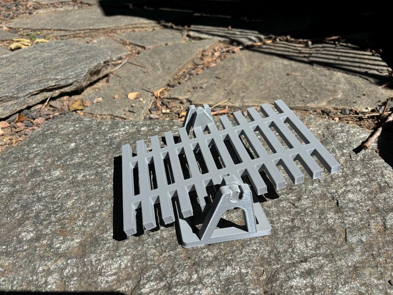 K&ouml;nnte beinhalten: Ein graues, 3D-gedrucktes Zaunelement mit zwei St&uuml;tzhaltern. Der Zaun hat eine Reihe von vertikalen St&auml;ben und ist so konzipiert, dass er aufrecht steht. Die Halterungen sind dreieckig und bieten Stabilit&auml;t. Der Artikel ist wahrscheinlich f&uuml;r ein Modell- oder Miniaturprojekt.