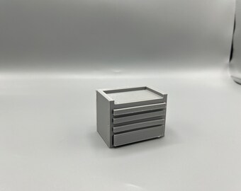 Könnte beinhalten: Eine kleine graue Metallwerkzeugbox mit vier Schubladen. Die Werkzeugbox hat eine flache Oberseite und ist aus glattem, mattem Material gefertigt.