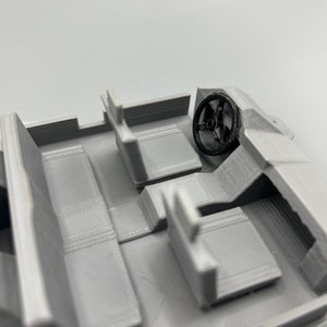 May include: A 3D-printed model of a vehicle interior, featuring seats, a dashboard, and a steering wheel. The model is primarily light gray with a black steering wheel, showcasing the interior's layout and design.