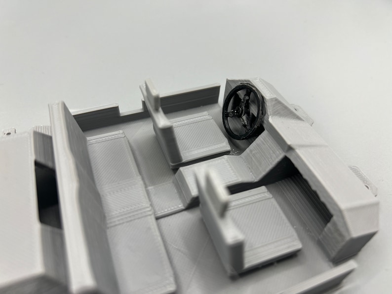 May include: A 3D-printed model of a vehicle interior, featuring seats, a dashboard, and a steering wheel. The model is primarily light gray with a black steering wheel, showcasing the interior's layout and design.