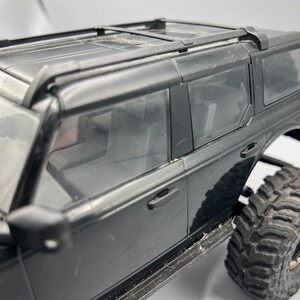 May include: Black toy SUV with large, rugged tires. The vehicle has a roof rack and tinted windows. The body of the SUV is black, and the tires are black with a gold rim. The vehicle is a model or toy.