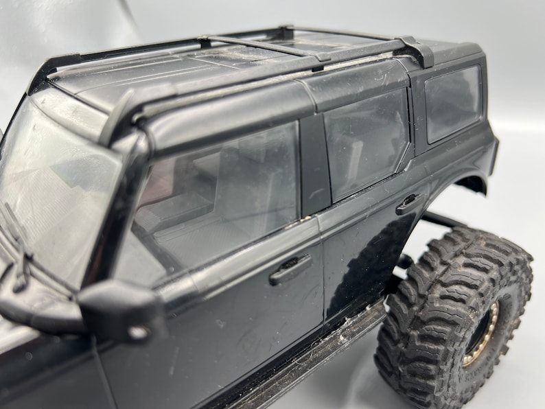 May include: Black toy SUV with large, rugged tires. The vehicle has a roof rack and tinted windows. The body of the SUV is black, and the tires are black with a gold rim. The vehicle is a model or toy.