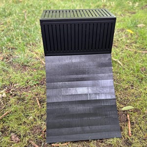 May include: A black knife block with a slatted top and a hinged, textured base. The base is angled, displaying a wood-grain pattern. The knife block is set on a bed of green grass.