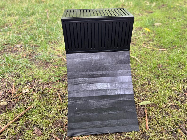 May include: A black knife block with a slatted top and a hinged, textured base. The base is angled, displaying a wood-grain pattern. The knife block is set on a bed of green grass.